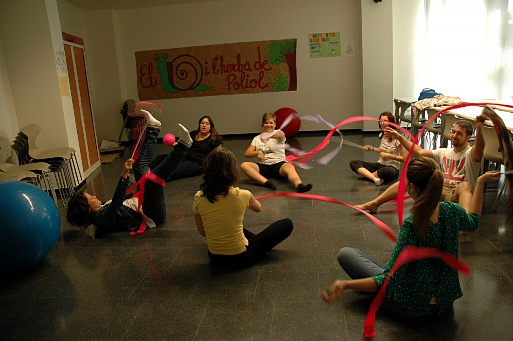 Estudiants dels graus d'Educació Infantil i Primària, asseguts a terra movent amb la mà unes cintes de colors